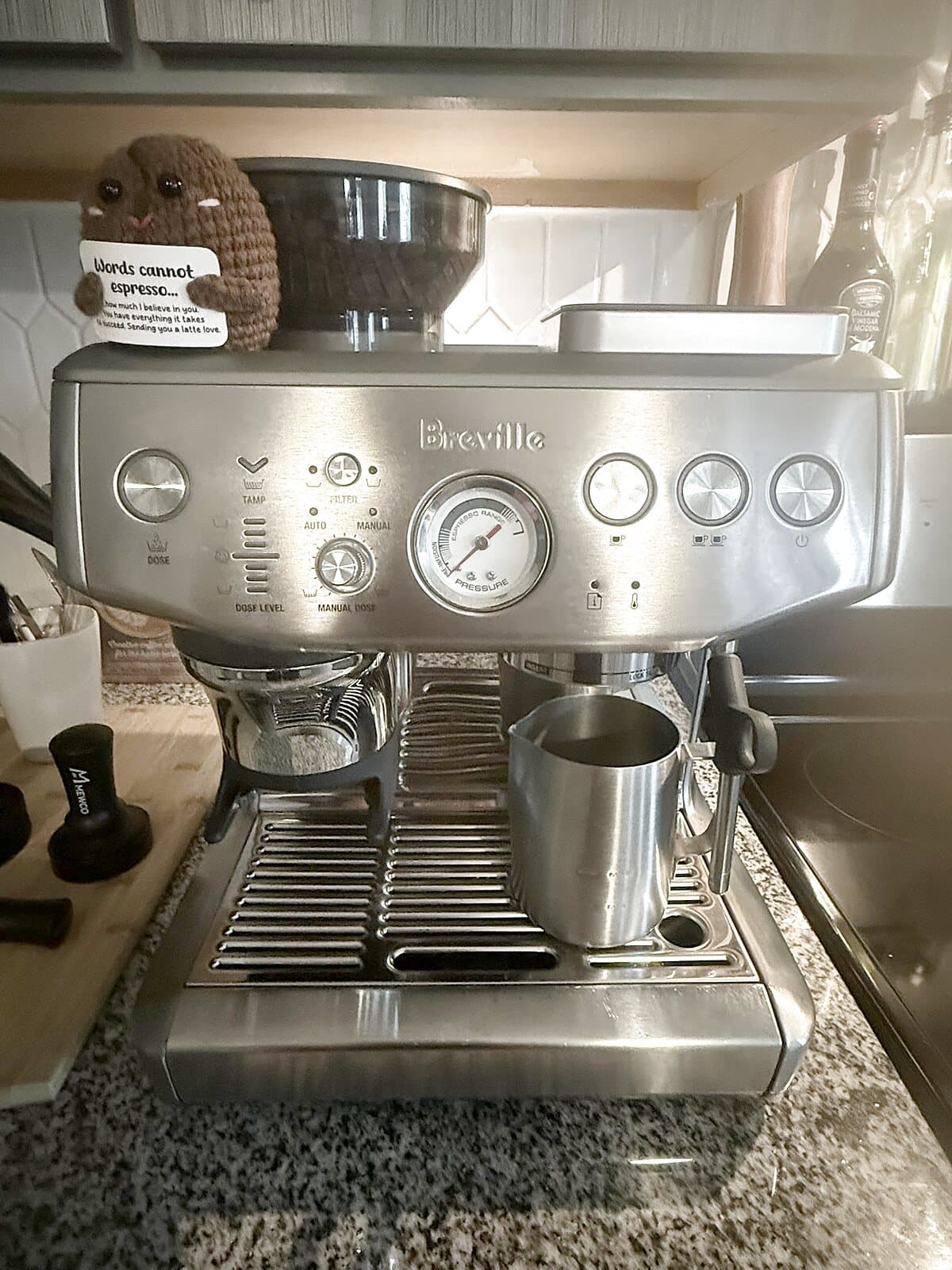 Front of the Breville Barista Express Impress with a steel milk pitcher on the drip tray and a MEWCO tamper beside it